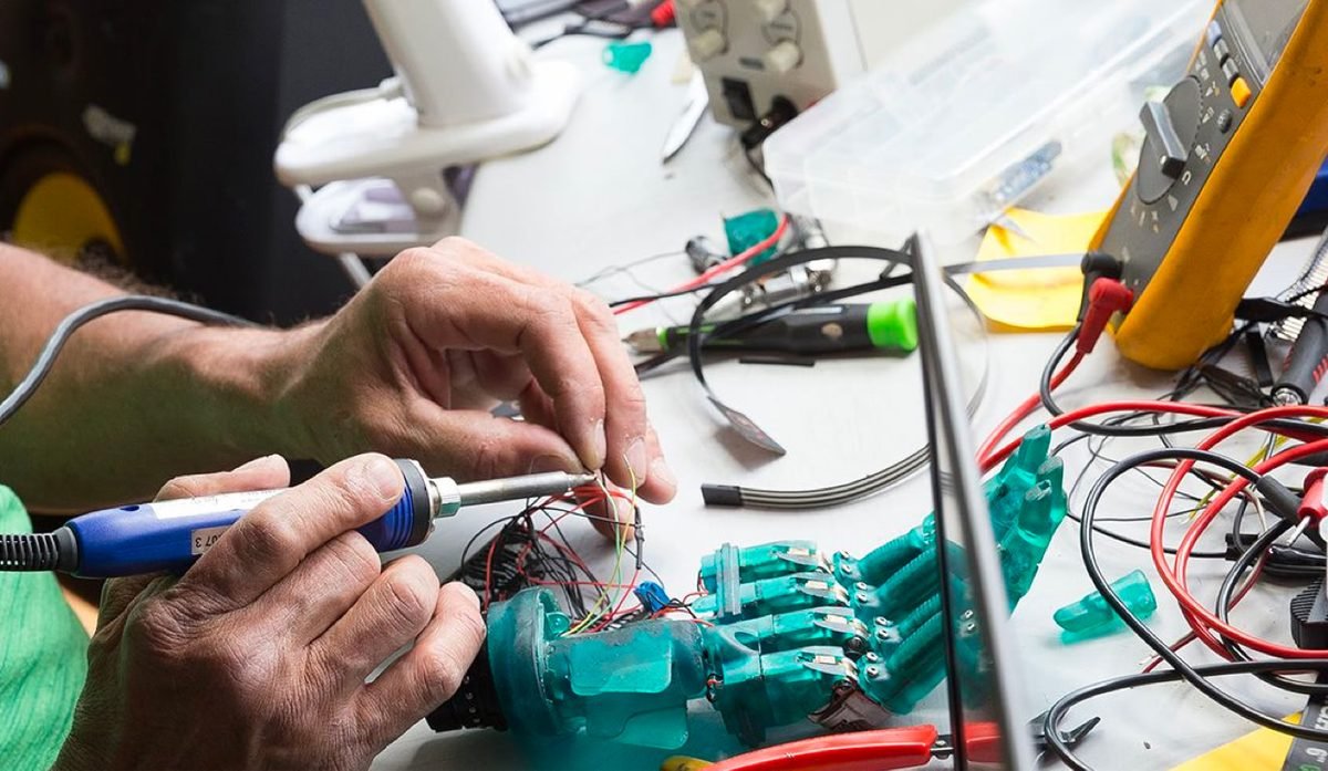 Manos soldando cableado en una pieza impresa en un banco de electrónica, con un multímetro y una fuente de poder detrás