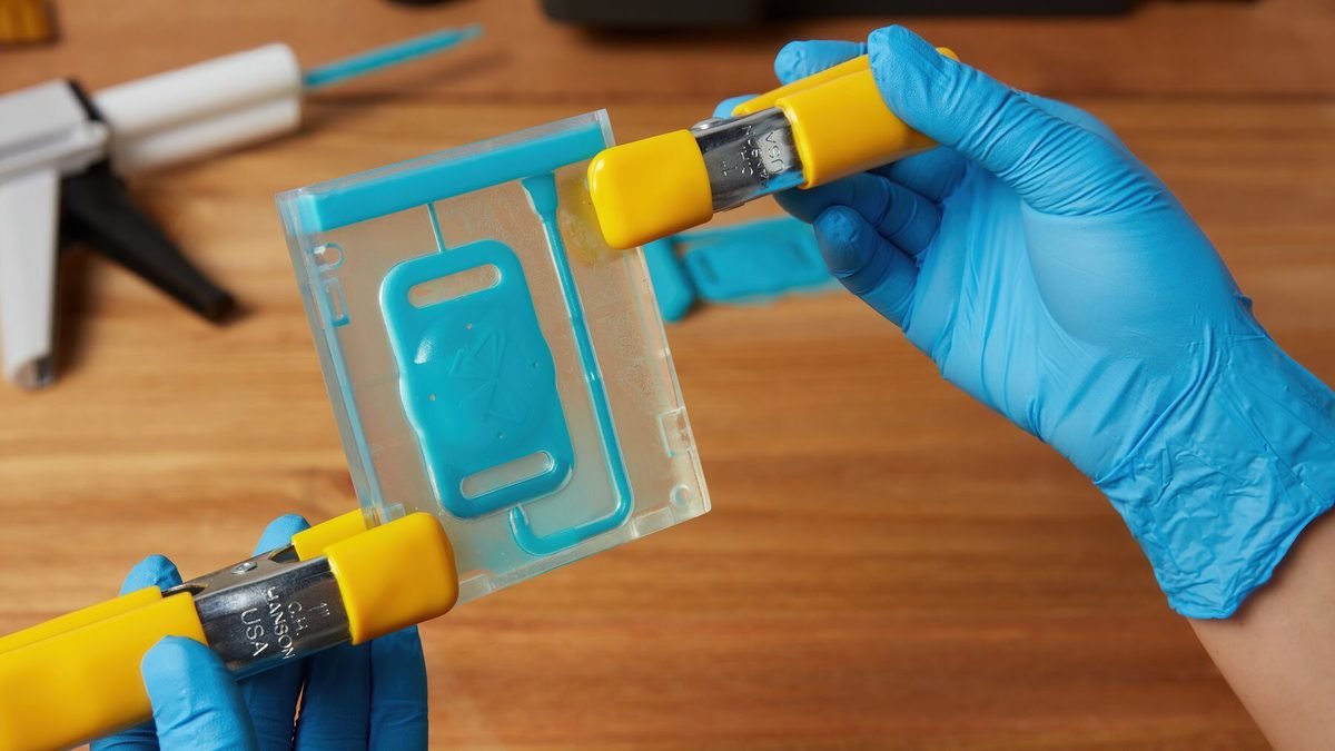 Gloved hands holding a clear printed part beside a yellow instrument